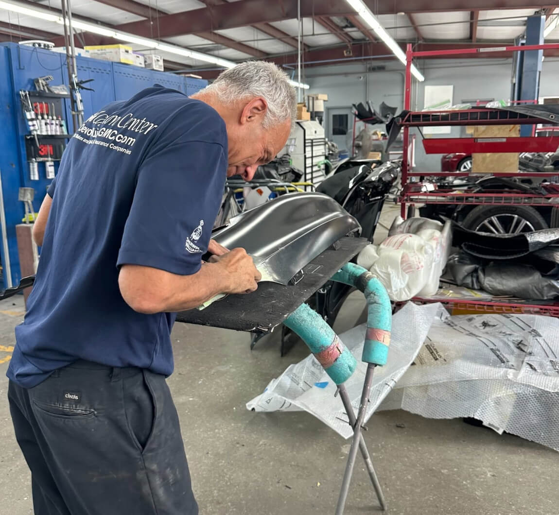 Side profile view of a collision center technician carefully hand-sanding a metallic gray vehicle bumper.