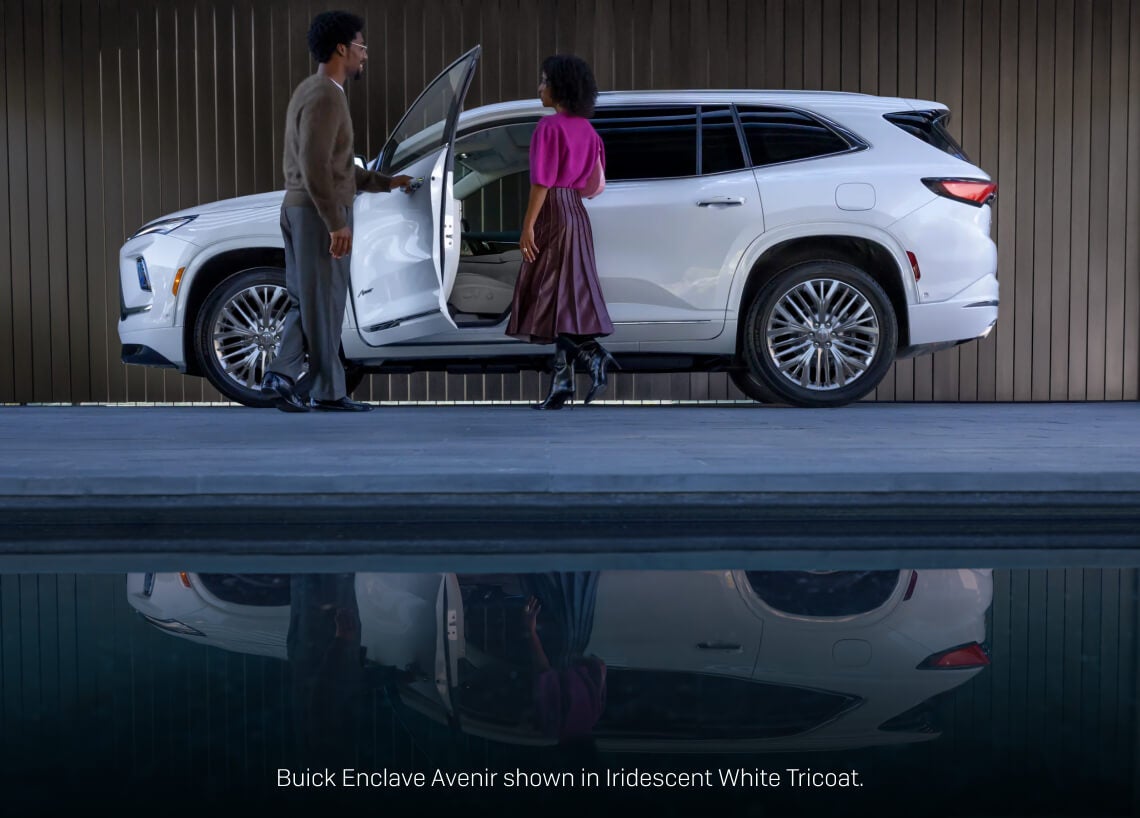Left profile view of an Iridescent White Buick Enclave Avenir reflected in a pool as a man opens the door for a woman.