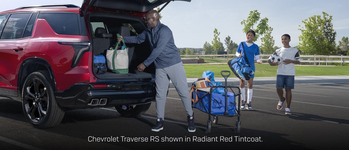 Rear three-quarter view of a Radiant Red Chevrolet Traverse RS as a family unloads sports gear in a park parking lot.