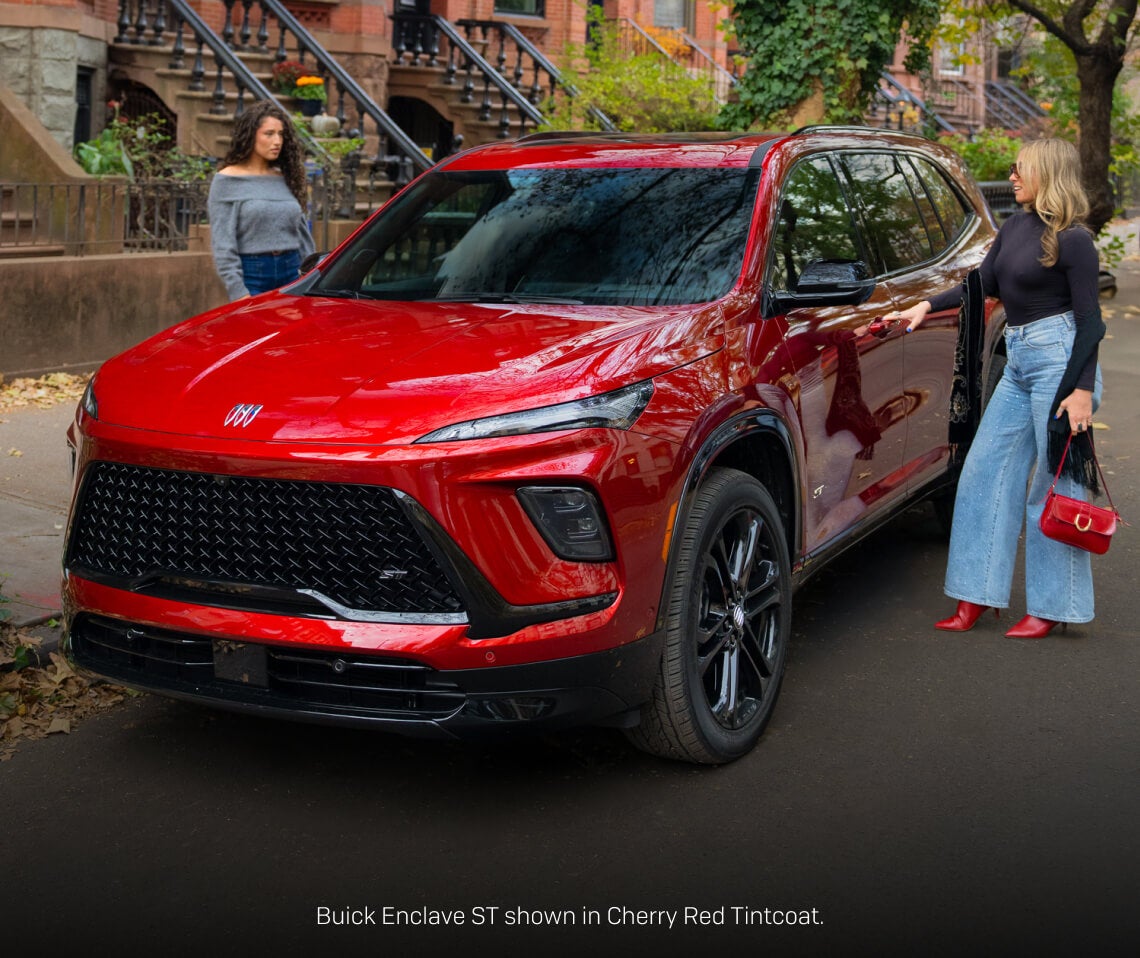 Front three-quarter view of a Cherry Red Buick Enclave ST parked curbside along a brownstone neighborhood street with two women.