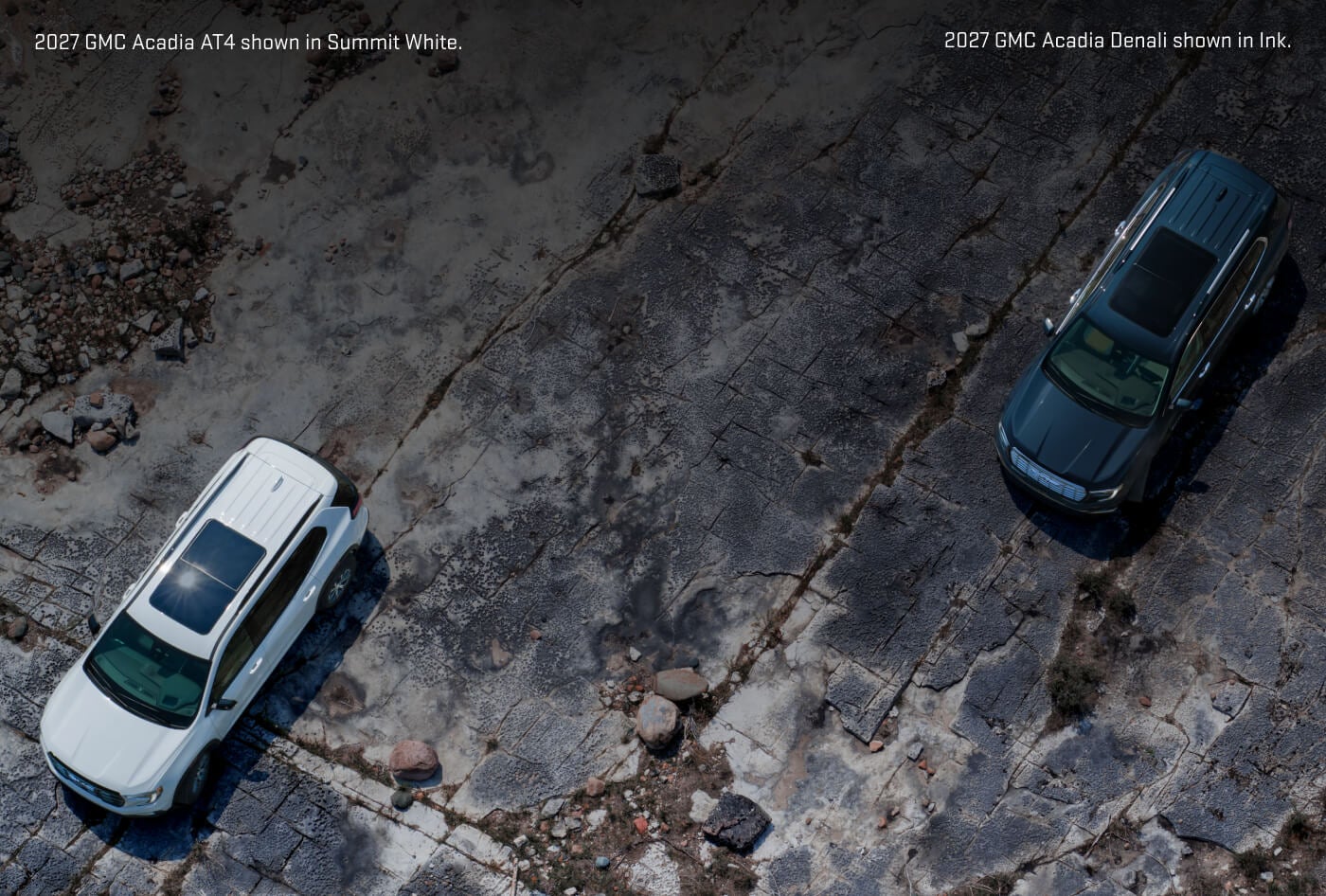 Aerial top-down view of a 2027 GMC Acadia AT4 in Summit White and an 2027 GMC Acadia Denali in Ink parked on rocky terrain.