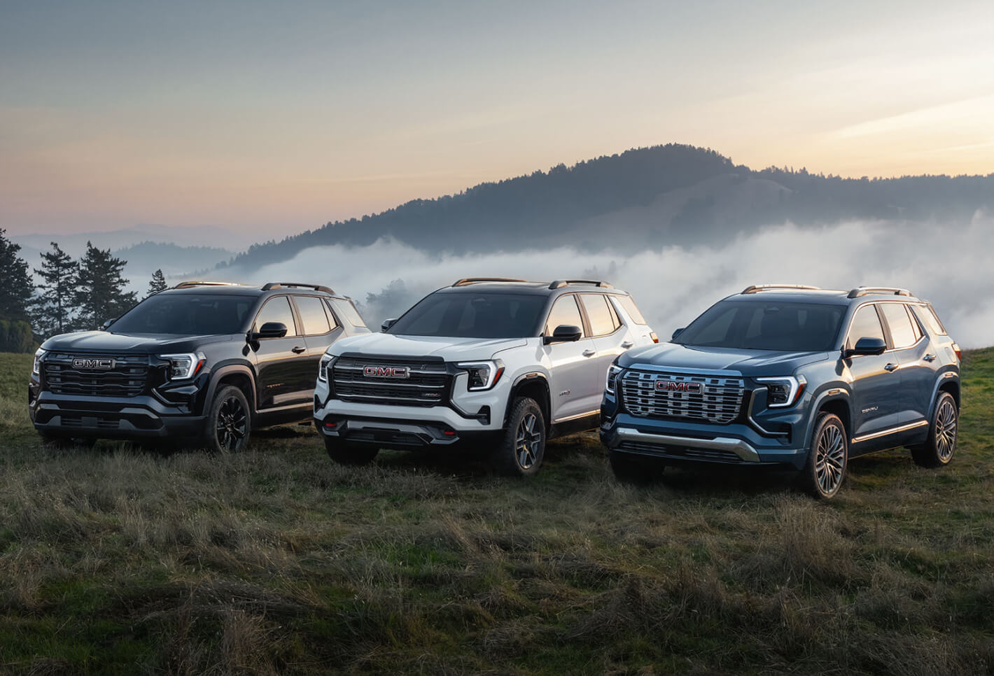 Front three-quarter view of 2027 GMC Terrain Elevation, AT4, and Denali trims in black, white, and blue on a misty hill.