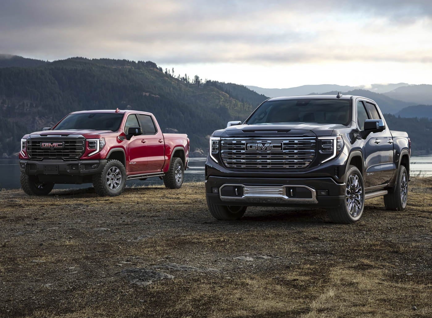 Two GMC trucks, one red and one black, are parked on rugged terrain near a lake.