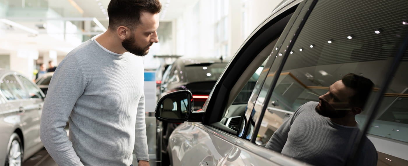 A man in a gray sweater closely examines a car's tinted window in a bright showroom.
